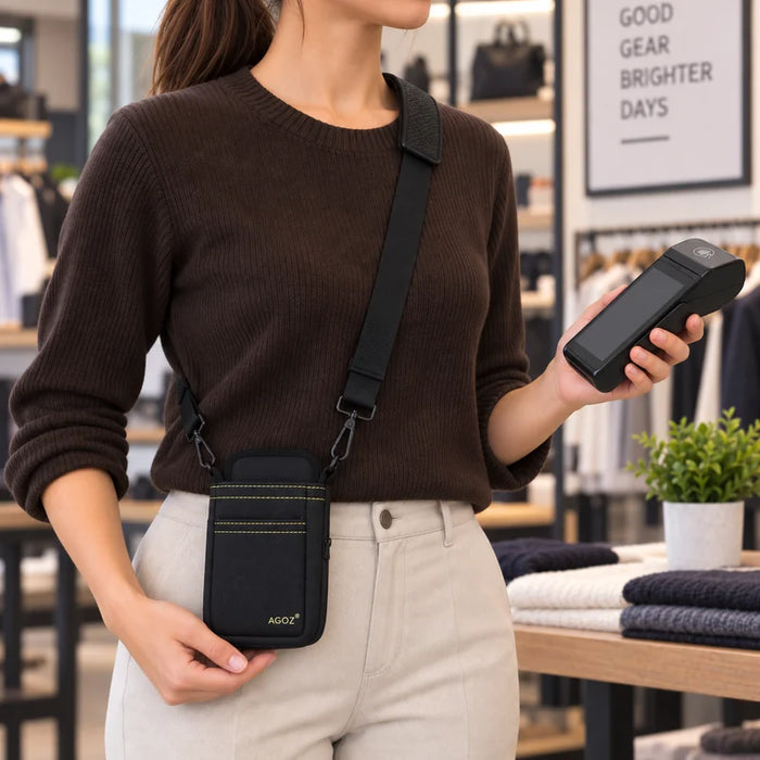 A woman wearing a brown ribbed sweater and beige pants models a black crossbody Adyen POS pouch with a black strap and subtle gold stitching in a retail store setting.