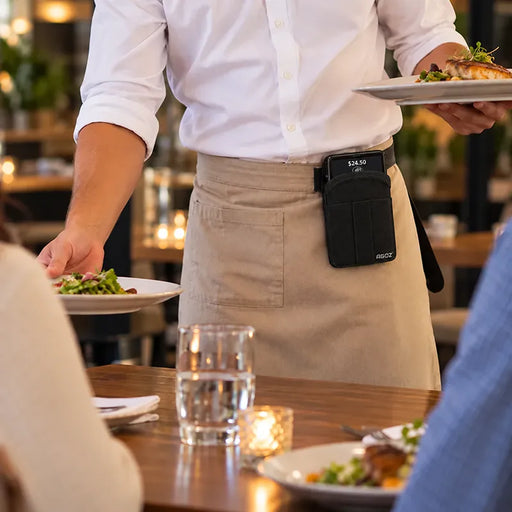 Restaurant server wearing the black Agoz Dejavoo POS holster clipped at the waist while carrying a plated meal in a dining setting.