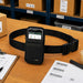 Black Newland mobile terminal waist belt holster displayed on a warehouse pick station with a Newland device, pick list, barcode labels, and packing materials.